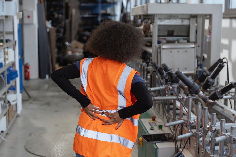Factory worker in a safety vest holding her lower back in pain while working near machinery