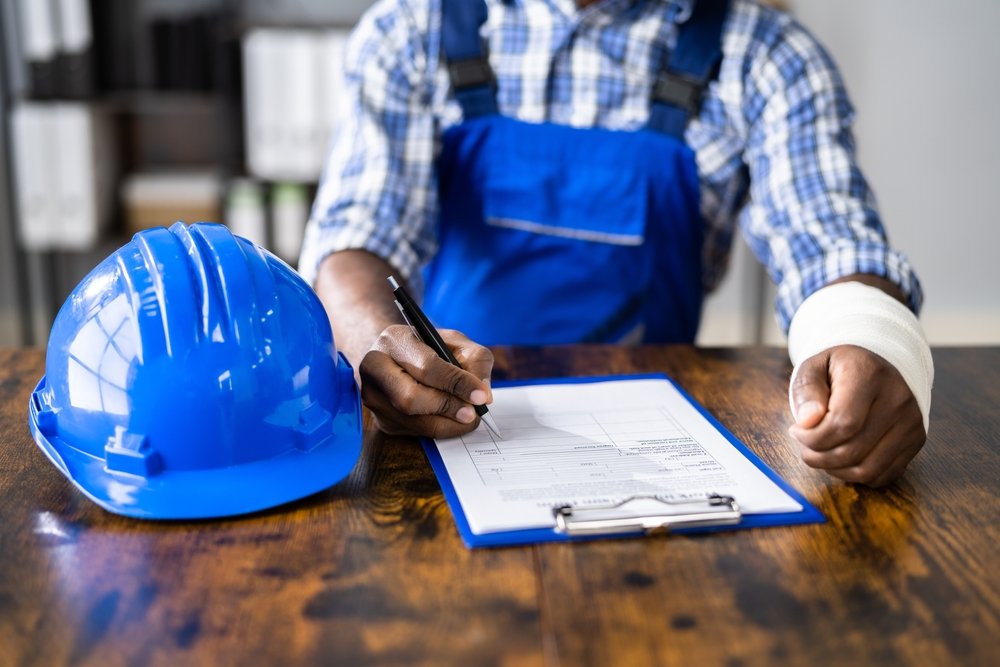 Construction worker with a bandaged wrist signing a workers compensation claim form beside a hard hat
