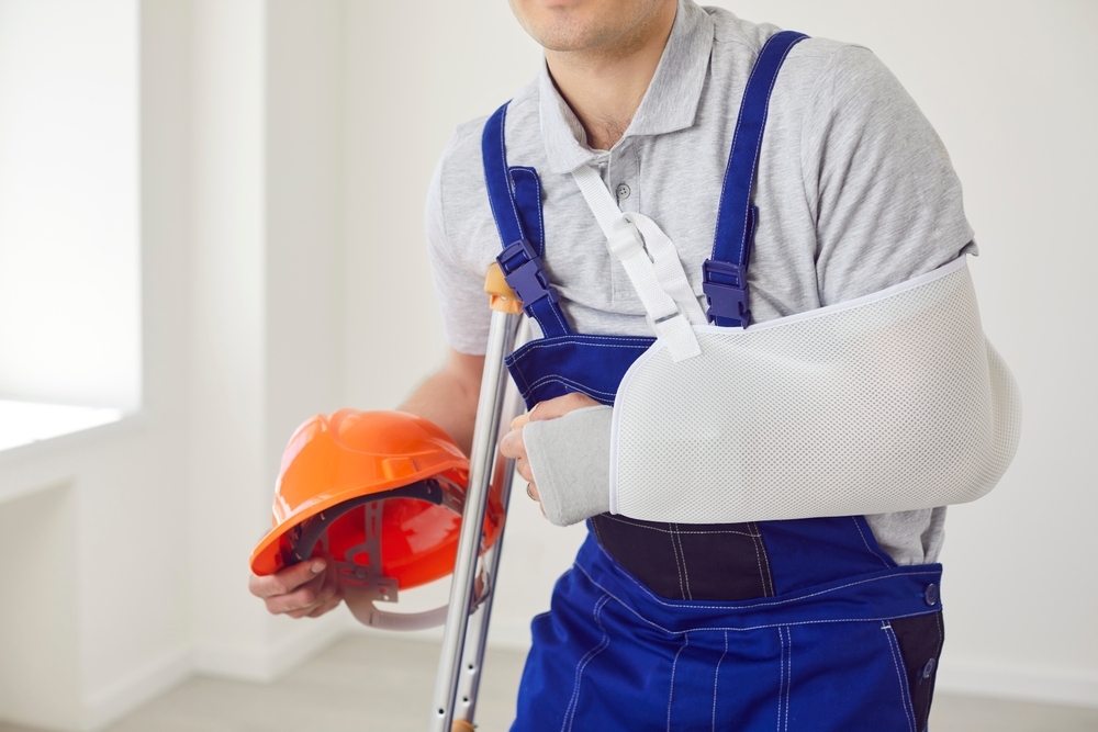 Construction worker with an injured arm in a sling holding a hard hat and crutch after a workplace injury Image 2 &mdash; worker with bandaged arm filling out injury cl
